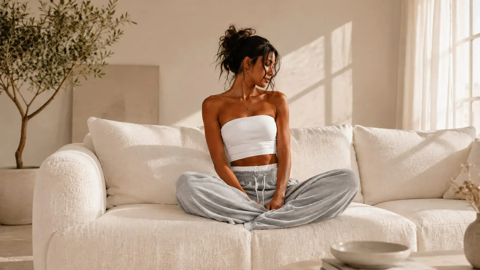 A woman sitting cross-legged on a cream boucle sofa in grey wide-leg sweatpants and a white top, lit by soft window light.