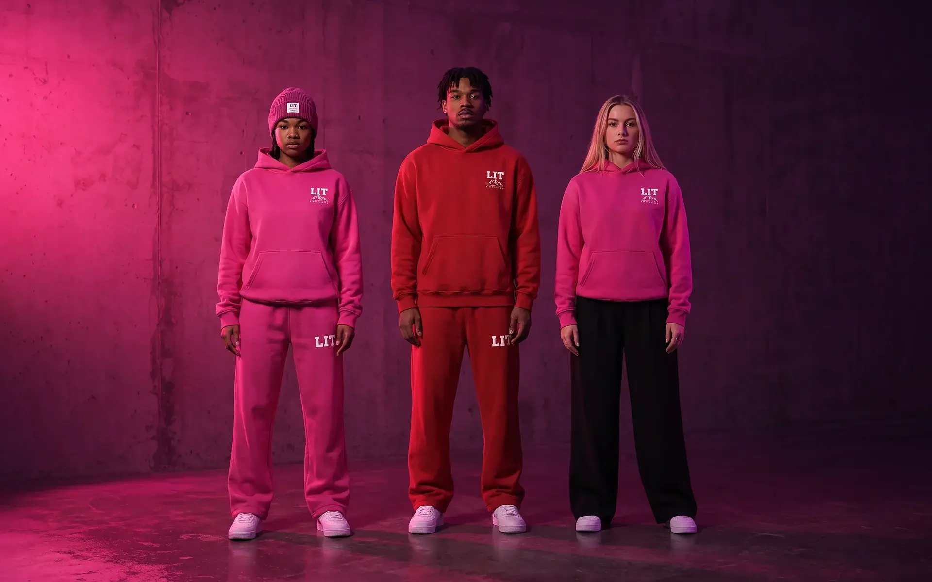 Three models in a concrete room under one magenta-gelled lamp: one in the pink LIT Sweat Suit and a fuchsia beanie, one in the red LIT Hoodie and Sweats, one in the pink LIT Hoodie.