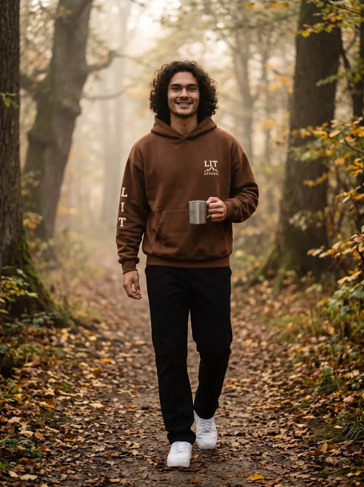 A smiling man walking a misty autumn woodland path in the chocolate LIT Sleeve Hoodie, holding a metal mug.