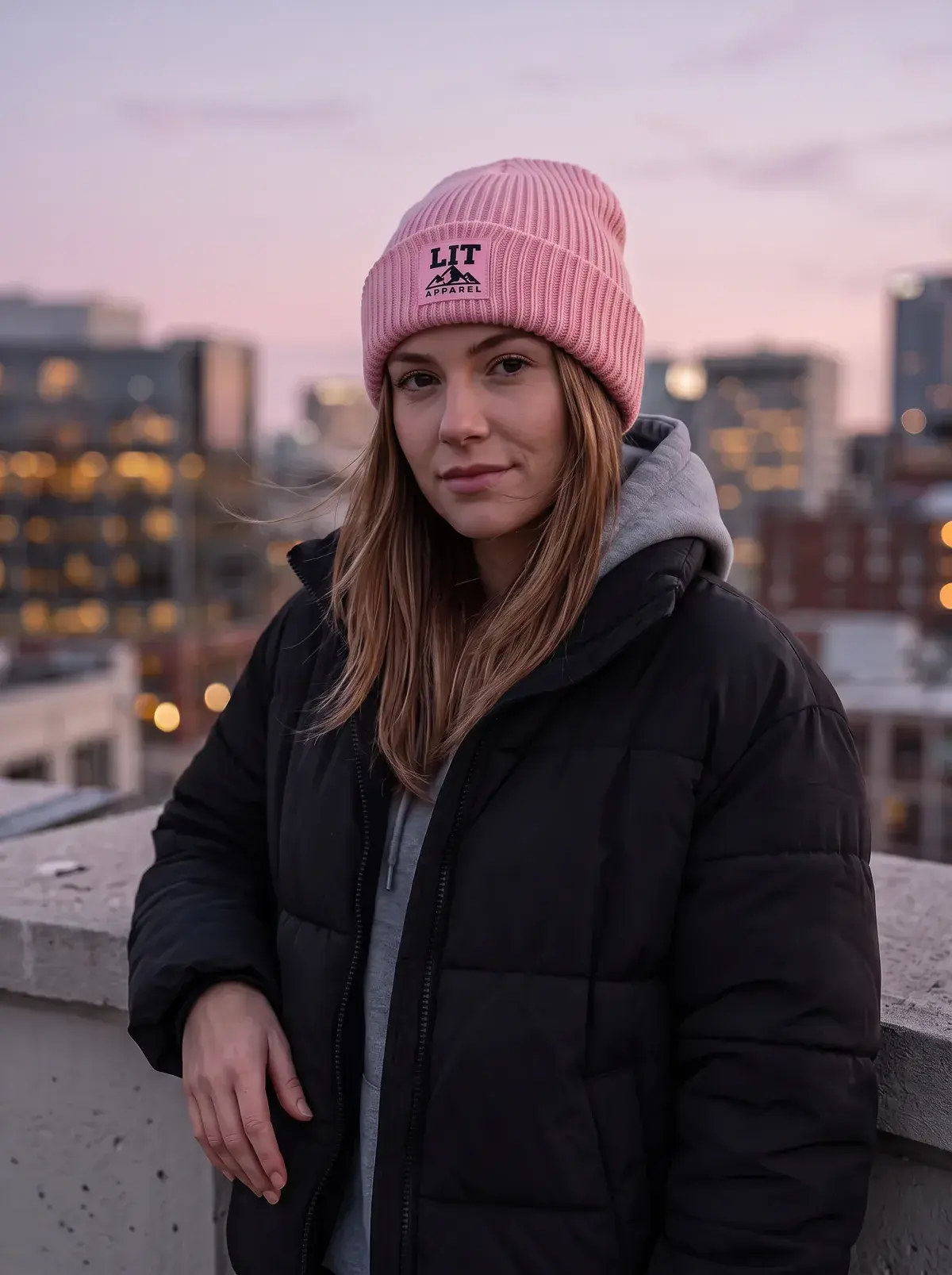 A woman in a pink ribbed LIT beanie and a black puffer on a rooftop at dusk, the city lit up behind her.